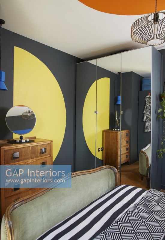 Bold and colourful painted bauhaus shapes in a contemporary bedroom. Showing yellow semi circle, vintage chest of drawers and a mirrored wardrobe. 