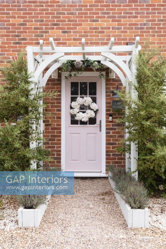 White pergola with shrubs over a pink front door with white floral wreath