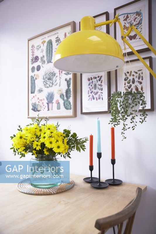 Dining table detail showing a vase of flowers, candles, a yellow floor lamp with botanical prints on the wall.