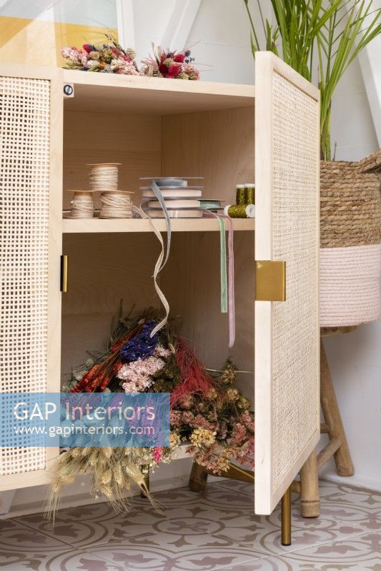 Freestanding wood craft cupboard with rattan doors storing ribbons and dried flowers 
