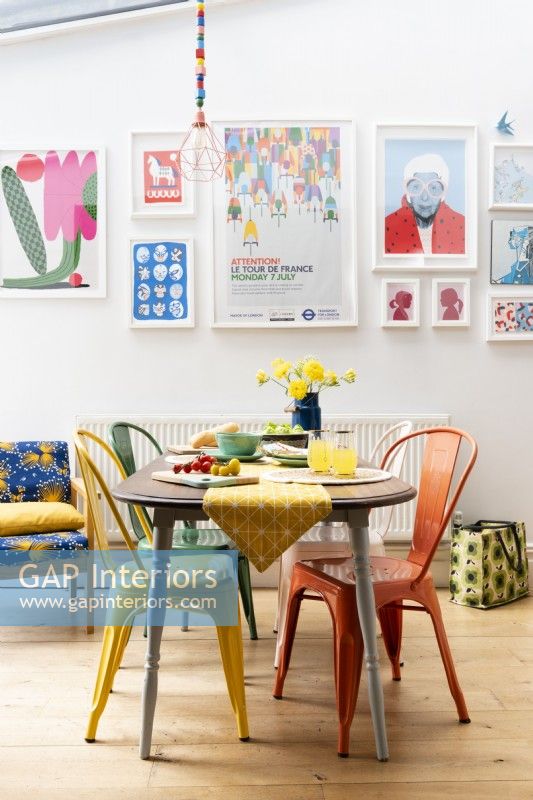 Reclaimed dining table with different coloured metal chairs in front of a colourful wall display of framed art