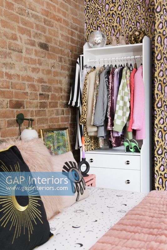 A white grab and go open wardrobe and drawer unit in a corner alcove of a bedroom against a wall of exposed brickwork and leopard print wallpaper