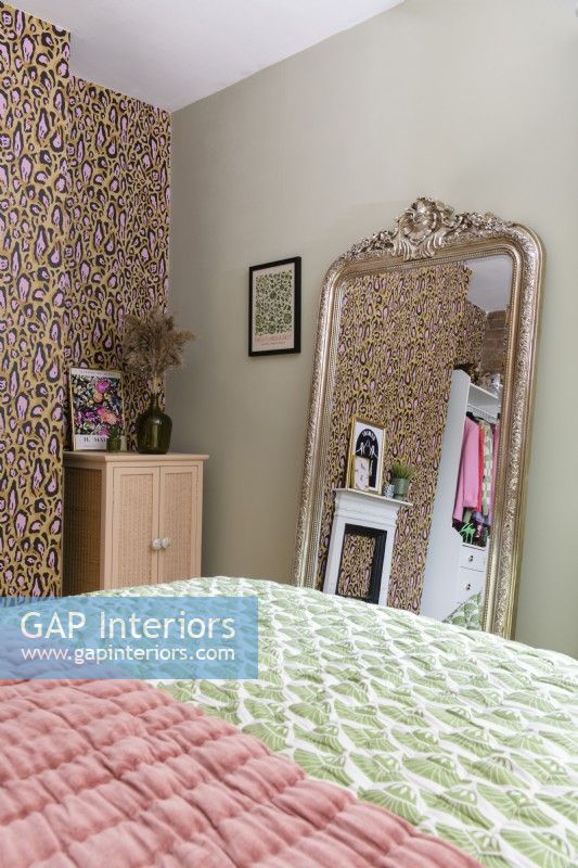 A large ornate gold framed mirror at the foot of a bed leans against a pale green wall in a bedroom with a feature wall papered with a leopard print design.