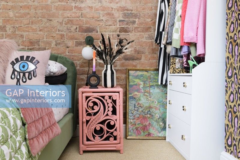 An exposed brick wall in a bedroom with a pink painted rattan bedside table next to a green velvet bed and an open grab and go wardrobe and drawer unit.