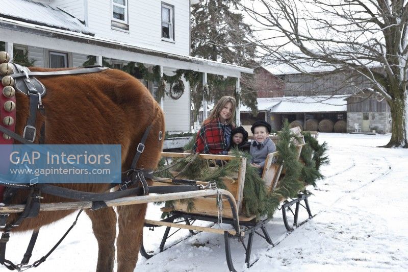 Mother and children going for a sleigh ride with partial views of the farmhouse and barn in the snow
