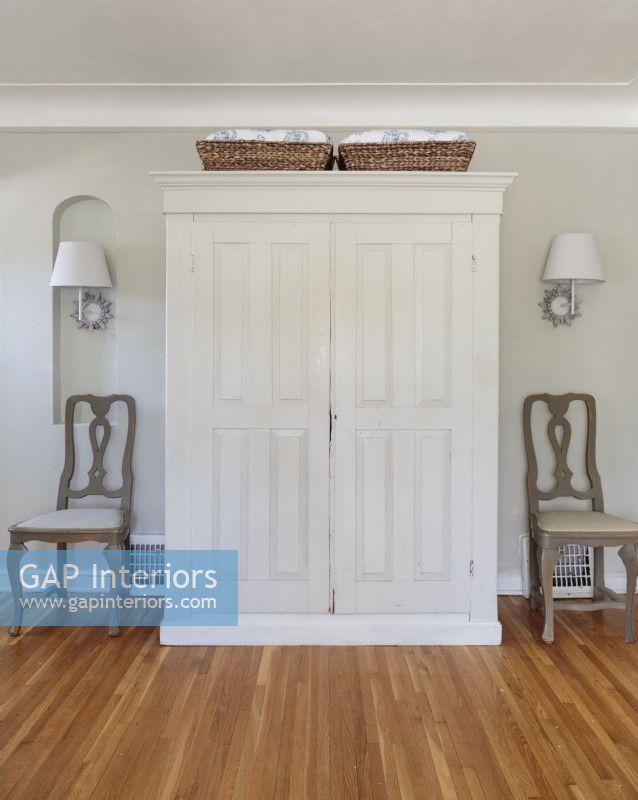 A vintage armoire got a makeover with white paint and is now used to store bed linens, blankets, throws and other necessary items. Symmetry is created with a pair of identical chairs, wall sconces and baskets.