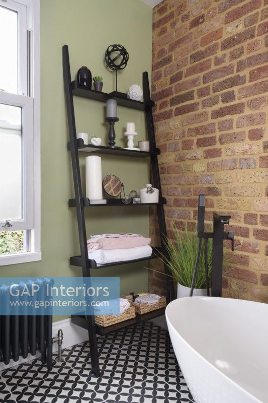 Black ladder shelving unit leaning against a green wall in the corner of a modern bathroom with an exposed brick wall