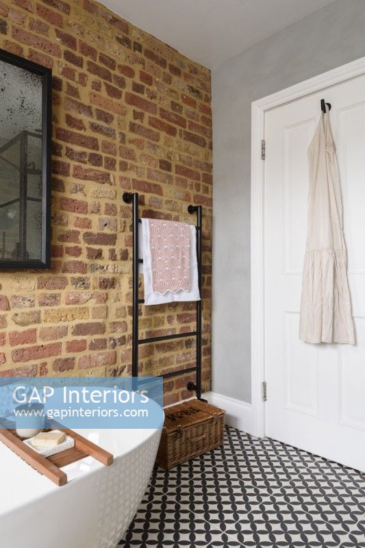 A black towel rail in the corner of a bathroom with exposed brickwork and a white door against a smooth grey cement wall with a monochrome patterned tiled floor