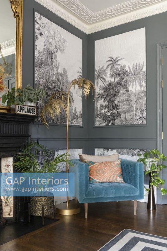 Gold fern floor lamp and a teal upholstered chair in the corner of a grey painted living room with paneling filled with monochrome tropical tree wallpaper