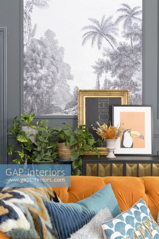 Detail behind an orange velvet sofa of a shelf with framed pictures and house plants in front of a black and white tropical tree patterned wall papered panel