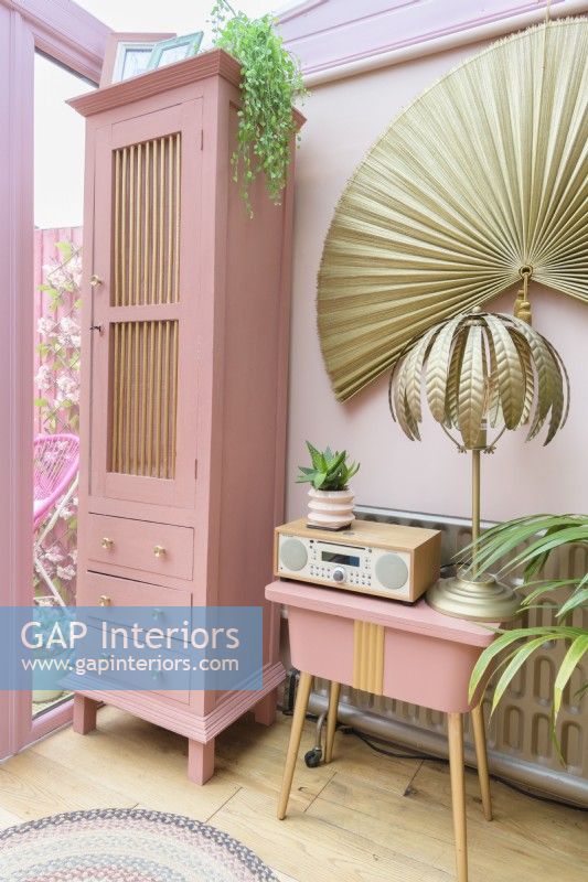 Tall pink upcycled cabinet and reclaimed pink painted vintage sewing table in a pink conservatory with gold tropical fern table lamp