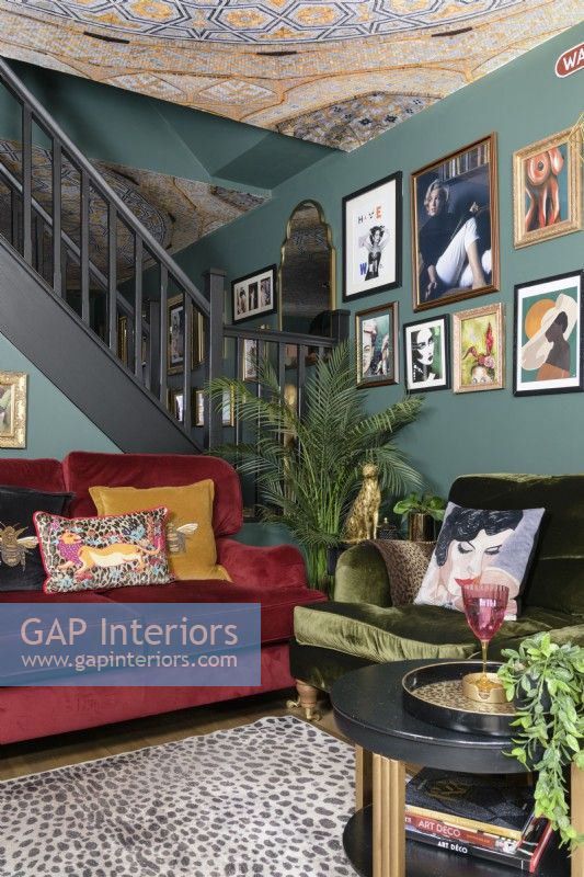 Open plan green living room with red and green velvet sofas in front of an open staircase and a salon style gallery art display wall and wall papered mosaic patterned ceiling
