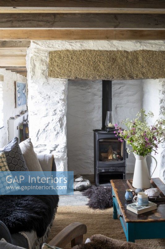 Living room with white washed walls, wooden beams and comfortable furniture.  Large vase of wild flowers picked from the surrounding countryside, and wood burner behind.