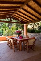 Villa Christina,  Kaminaki, Corfu, Greece. Covered outdoor terrace dining area with wooden table and chairs set for alfresco dining