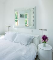 Modern white bedroom with mirror above bed and bedside table with vase of stocks