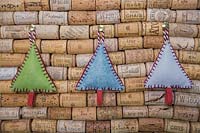Making stitched felt christmas decorations - miniature christmas trees made from felt and decorative string, hanging against a wine cork board