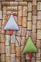 Making stitched felt christmas decorations - miniature christmas trees made from felt and decorative string, hanging against a wine cork board