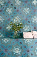Colourful tiled wall in bathroom  with towels and vase of flowers 
