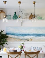 Coastal painting and shelf of glassware above dining table 