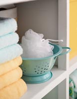 Miniature colander with exfoliating cloth and towels on bathroom shelf