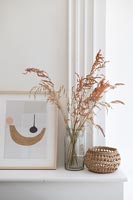 Cut grasses in glass jar with wicker basket and artwork on mantelpiece 
