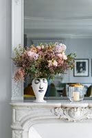 Vase face painted on it containing flower arrangement on mantelpiece 