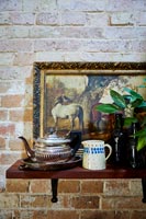 Silver teapot on shelf with painting against exposed brick wall 