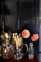 Cutlery in glass jugs next to dahlias in vase against black wall 