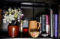 Flowers in ceramic jug next to books and ornaments on black shelf 