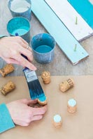 Woman painting tops of corks in various shades of turqouise using a paintbrush