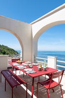 Red dining table and chairs on white terrace with sea views in summer 