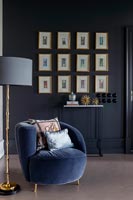 Blue armchair with floor lamp and display of framed pictures on black wall 