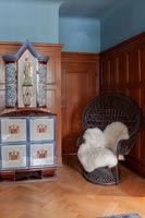 Large high backed rattan chair next to unusual cabinet in panelled hallway