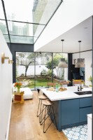 Contemporary modern kitchen in a side return extension with a blue island, wooden floors, light pendants, bar stools and slim frame tall aluminium doors.