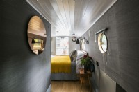 Contemporary bedroom on canal, narrow, river boat with dark grey walls, wooden floors, white painted ceiling, round mirror, porthole window and yellow throw.