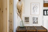 Bright white upstairs hallway with built-in storage and cupboards. Framed pictures hang on the walls. In the foreground a steamer trunk stands with a vase with pampus grass. 