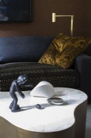 Figurine and sculpture displayed on white marble and brass coffee table in front of dark blue sofa and gold pillow.
