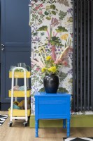 Upcycled painted cabinet table against floral wallpaper with yellow storage trolley