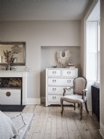 Classic bedroom featuring upholstered chair, chest of drawers and former fireplace