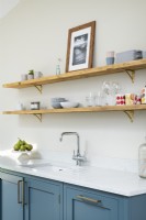 Open wooden shelves in modern classic kitchen with brass brackets, blue units, white countertop and taps.