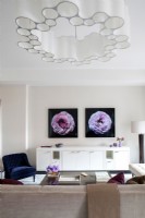 Modern living room with long white credenza decorated with photographs of flowers on wall and unique chandelier.