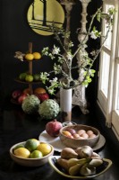 Bowls of fruit next to branches with blossom in vase against black wall
