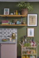 Kitchen detail with a gold kettle, wooden open shelving, green painted walls, patterned tiles and a drinks trolley.