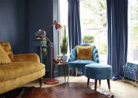 Living room showing a mustard yellow sofa, turquoise armchair with footstool, navy painted wall and a floor lamp. 