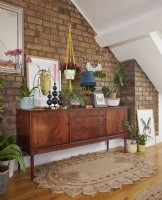 Attic living room with a mid century sideboard with plants, candles and artwork against an exposed brick wall.