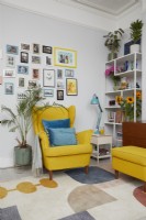 Living room corner showing yellow mid century style armchair and footstool with a gallery wall, shelf unit and plants.