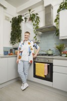 Woman in a kitchen with blue metro tiles, hanging plants and a green saucepan.