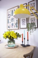 Dining table detail showing a vase of flowers, candles, a yellow floor lamp with botanical prints on the wall.