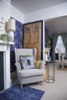 Living room with a cosy armchair, blue patterned wallpaper and a view through to the dining room.