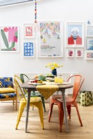Reclaimed dining table with different coloured metal chairs in front of a colourful wall display of framed art